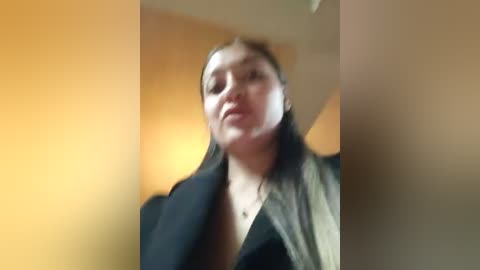 A blurred, grainy video of a woman with light skin and brown hair, wearing a black jacket, standing against a beige wall. Her expression is neutral.