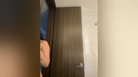 Video of a person with light skin, visible tattoos, and a toned arm partially blocking a wooden door with a metallic handle, in a modern, minimalist room with white walls.