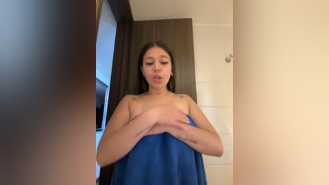 Media: A video of a young Asian woman with straight black hair, medium build, and small breasts, wearing a blue towel, standing in a modern, beige-tiled bathroom.