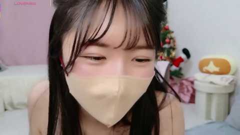 Media: A close-up video of an Asian woman with long black hair, wearing a beige surgical mask, in a softly lit bedroom with a Christmas tree and a plush toy.