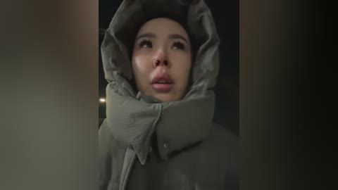 Media: Video of a young woman with fair skin, light brown hair, and wide eyes, wearing a green puffy jacket with fur lining, looking upwards with a contemplative expression.