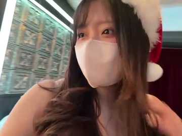 Video of an Asian woman with long, dark hair wearing a red and white Santa hat, white mask, and a sleeveless top, standing in a brightly lit room with a blurred background of a currency display.