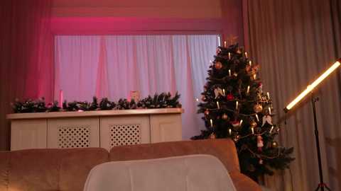 Video of a cozy living room with a lit Christmas tree, festive garland, soft pink and white lights, beige sofa, and white curtains.