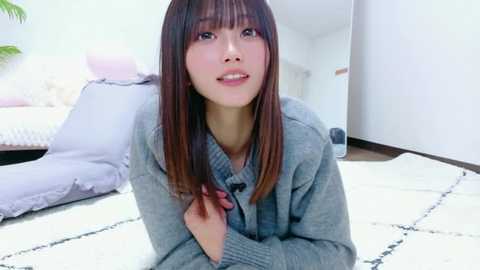 Video of an Asian woman with straight, shoulder-length brown hair, wearing a gray sweater, sitting on a white rug in a modern, minimalist living room with white walls and a purple sofa.