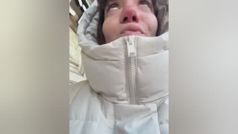 A close-up video of a young woman with fair skin and brown hair, wearing a puffy, white winter jacket, looking upward with a thoughtful expression.