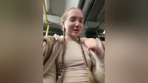 Media: A video of a smiling young woman with light skin and braided hair, wearing a beige jacket and shirt, seated on a yellow bus.