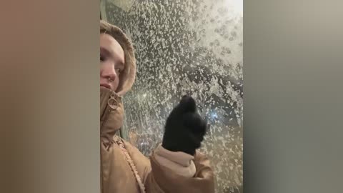 Video of a person in a tan winter coat, standing indoors, looking through a frosted glass door. Snowflakes are visible on the glass, creating a wintery ambiance.