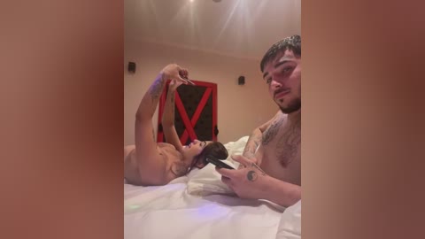 Video of a man with tattoos, lying on a bed, holding a black dildo to a woman's mouth, who is tied up with red rope, in a dimly lit room.