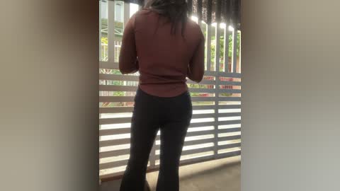 A video of a woman with long, dark hair wearing a maroon top and black pants standing at a railing, looking out at greenery and a white fence.