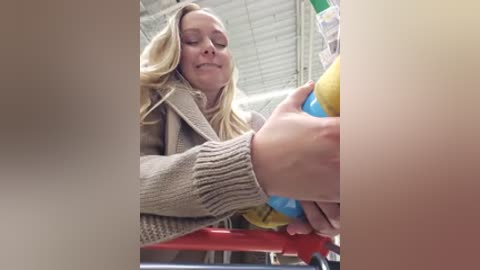 Video of a smiling blonde woman in a beige sweater, holding a blue and yellow ball, in a brightly lit, industrial warehouse with metal beams and shelves.