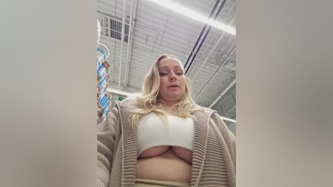 Media: Video of a blonde woman with a curvy physique, wearing a white crop top and beige cardigan, taken from a low angle, showing her cleavage, in an industrial setting with metal beams and ventilation.