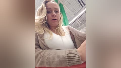 Media: Video of a blonde woman in a beige cardigan over a white top, seated indoors with green curtains and industrial ceiling.