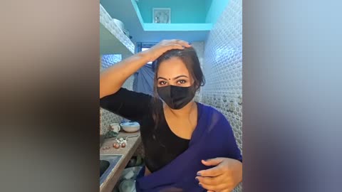 Video of a woman with medium brown skin, dark hair, and a black face mask, seated in a small, tiled bathroom with a mirror. She wears a black top and blue shawl, and appears to be applying makeup.