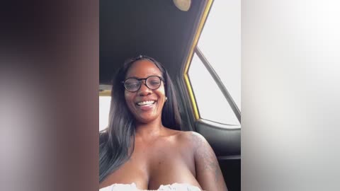 Video of a smiling Black woman with long braided hair, wearing glasses and a white lace bra, seated in a car with sunlight streaming through the window.