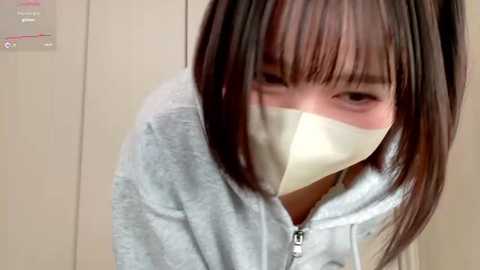 A video of an East Asian woman with straight, shoulder-length black hair, wearing a light gray hoodie and a beige surgical mask, looking down slightly with a neutral expression.