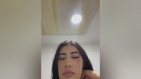 Media: A close-up video of a Latina woman with long black hair, wearing makeup, including dark eyeliner, pink blush, and nude lipstick. She is smiling with her eyes closed, captured from a low angle. The background is a white ceiling with a single bright light.