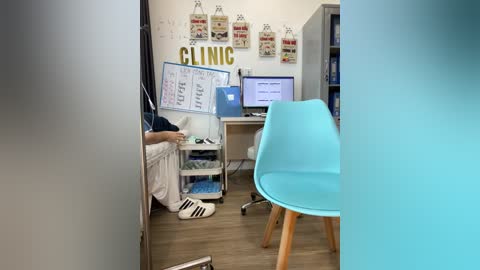 Media: Video of a small office with a person sitting at a desk, wearing a white shirt and black pants, surrounded by cluttered shelves, a computer monitor, and a blue chair.