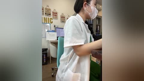 Video of an Asian woman with long black hair in a white lab coat and mask, working at a desk in a bright office with framed signs, computers, and a green chair.