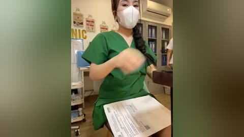 Video of a female nurse in green scrubs, face mask, holding papers, in a hospital room with a wall sign, cabinets, and a bed.