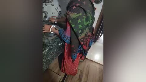 Media: A video of a person with dark skin, wearing a patterned headscarf and a red dress, sitting at a kitchen counter with a white wristband. The background includes kitchen appliances and a wooden floor.