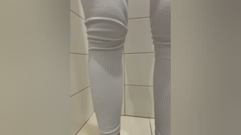 Video of a person's legs in white ribbed leggings, standing in a tiled bathroom with beige walls and white floor tiles.