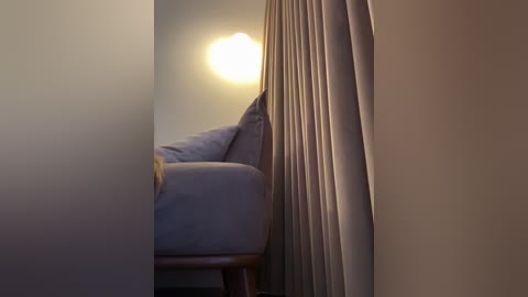 Media: Video of a cozy, dimly lit bedroom corner with a beige curtain partially drawn, revealing a portion of a light grey upholstered chair and a bed with white sheets. The room has soft, warm lighting, giving a serene and intimate atmosphere.