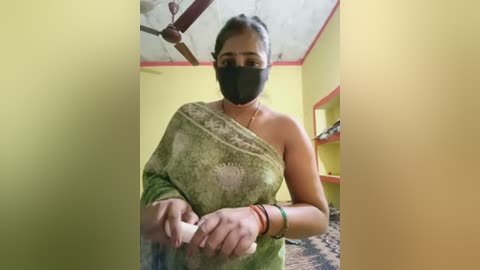 Media: Video of a woman in a green saree with a paisley design, wearing a black mask, standing in a yellow-painted room with a fan and shelves.
