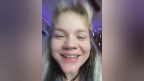 Video of a young, light-skinned woman with blonde hair, smiling broadly. She has a small nose stud and is wearing a light-colored top. The background is blurred, suggesting an indoor setting with purple lighting.