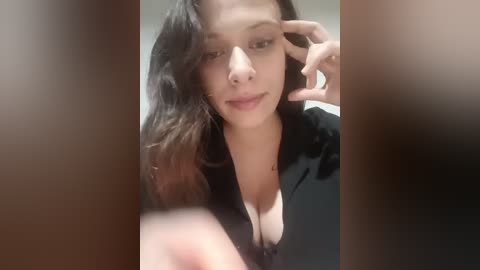 A video of a young woman with long, wavy brown hair, fair skin, and a black top, gazing intently with one hand touching her face. Background is blurred.