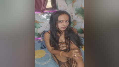 Video of a young, slender South Asian woman with long black hair, wearing a revealing black fishnet bodysuit, posing seductively on a bed with colorful bedding. The background features a wall with peeling paint and a window with pink curtains.