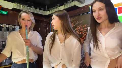 Media: Video of three young women with light skin, one with blonde hair, one with long brown hair, and one with long black hair, wearing white tops and drinking colorful smoothies in a cozy, dimly-lit cafe.