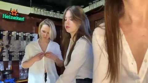 Media: Video of three young women in a dimly lit bar. One adjusts her white blouse, another stands with long hair, and a third is partially visible in the background.