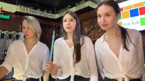 Media: Video of three young women with long hair, dressed in white shirts and dark pants, standing in a modern bar with colorful digital screens and glassware.