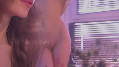 A close-up video of a nude woman with light brown skin and long wavy hair, seen from the side, with her breasts partially visible. Background features white blinds and a blurred indoor setting.