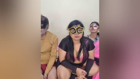 Video of three women in elaborate, revealing superhero costumes with black, pink, and yellow accents. They are seated in a plain room, with blurred figures in the background.