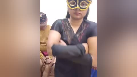 A video shows a woman with medium skin tone, wearing a black outfit, yellow mask, and black gloves, seated next to an older man in a yellow shirt and red pants.