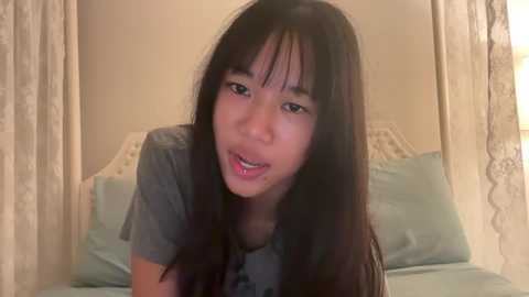 Video of a young Asian woman with long black hair and a slight smile, wearing a gray t-shirt, lying on a bed with light blue sheets and beige curtains in the background.