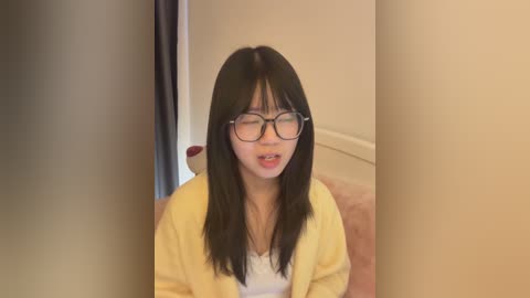 Video of an Asian woman with long black hair, wearing glasses, a yellow cardigan, and a white top, sitting in a beige room with a carpeted floor.