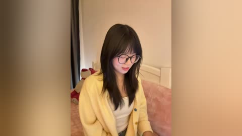 Video of an East Asian woman with long black hair, wearing glasses, a yellow cardigan, and a white top, sitting on a bed with pink sheets, in a softly lit room.