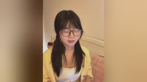 Video of an Asian woman with long black hair, wearing glasses and a yellow cardigan over a white top, standing in a beige room with a pink carpet.