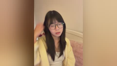 Video of an Asian woman with long black hair and glasses, wearing a yellow robe over a white top, sitting on a pink carpet in a beige room.