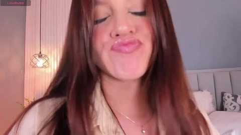 Video of a young woman with long brown hair, fair skin, and closed eyes, puckering her lips, wearing a beige top and gold necklace, in a cozy bedroom with a bed and modern pendant light.