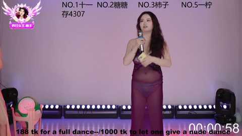 Video of a woman singing into a microphone on a stage, wearing a sheer, dark purple dress, with a watermark logo in the corner.