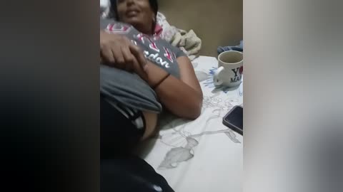 Media: Video of a woman with medium skin tone, wearing a gray t-shirt and black shorts, lying on a bed with a patterned white sheet. She holds a cup and a smartphone beside her.