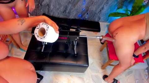 Video of two nude women with tan skin, one bent over a black leather bench, the other holding a white condom, in a modern, indoor setting with tiled floor and plants.