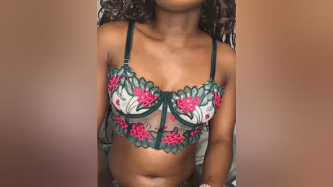 Video of a dark-skinned woman with curly hair, wearing a sheer, green lace bra with red floral embroidery, against a plain, light brown background.