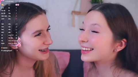 Video of two young Asian women with light skin, smiling and looking at each other, in a cozy indoor setting with a pink pillow.