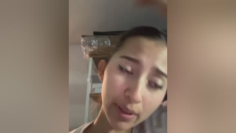 Media: Video of a young woman with light skin and dark hair, wearing a light-colored shirt, looking down and slightly frowning, against a blurred, beige wall background.