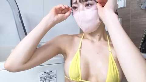 Media: Video of an East Asian woman in a yellow bikini, adjusting a pink face mask, in a modern bathroom with white walls and a glass shower.