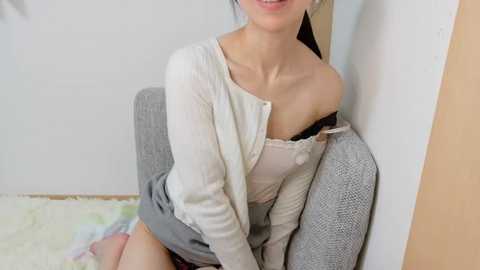 Media: A video of a smiling Asian woman with light skin, wearing a white cardigan, black bra, and gray skirt, sitting against a gray cushioned chair, in a simple room with white walls and wooden floors.